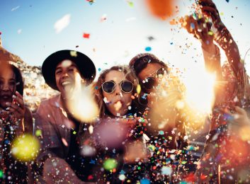 Group of teenager hipster friends partying by blowing and throwing colorful confetti from hands with sunset sun flare